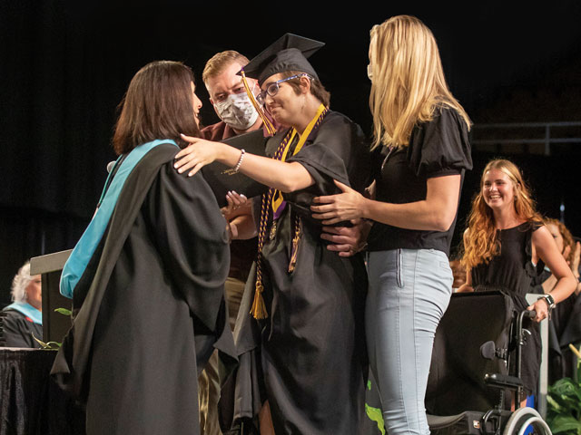 Hailey stands on the stage at her high school graduation and reaches out to hug her school principal. Hailey is smiling and wears a black graduation cap and gown, a medal, and purple and gold academic cords draped over her shoulders. Her school principal is also smiling and wears a black academic gown with a hood lined with light blue velvet. Two therapists stand to either side of Hailey, ready to help her if she needs assistance walking or standing. Behind the group is Hailey’s sister, who is smiling and holding a wheelchair.”