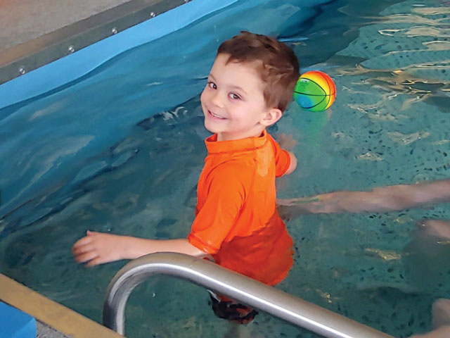 Tripp smiles during aquatic therapy. An inflatable ball floats in the pool behind him.