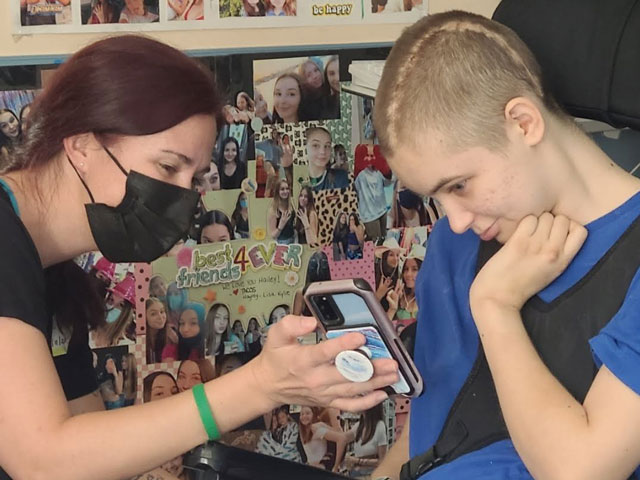 A woman holds up a phone for Hailey to look at. Hailey, shown in profile, is smiling and sitting in a wheelchair, supporting her head with her hand. Her hair is very short, and a large scar is visible from her forehead to the back of her neck. Behind them is a bulletin board with cards and photos of Hailey and her friends.