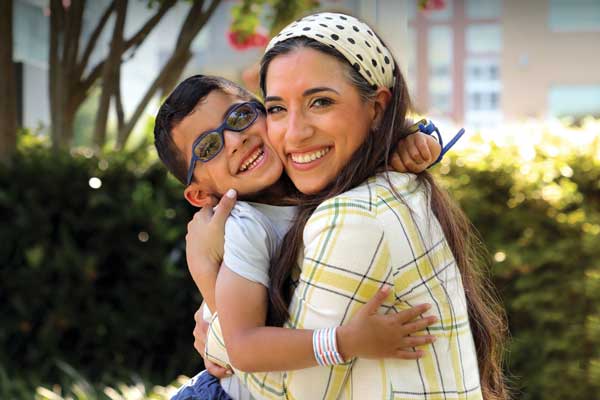 A young boy hugs his mom outside. Both have big smiles. The boy wears glasses and a kippa, and has a birthmark on his right ear and on the right side of his neck.