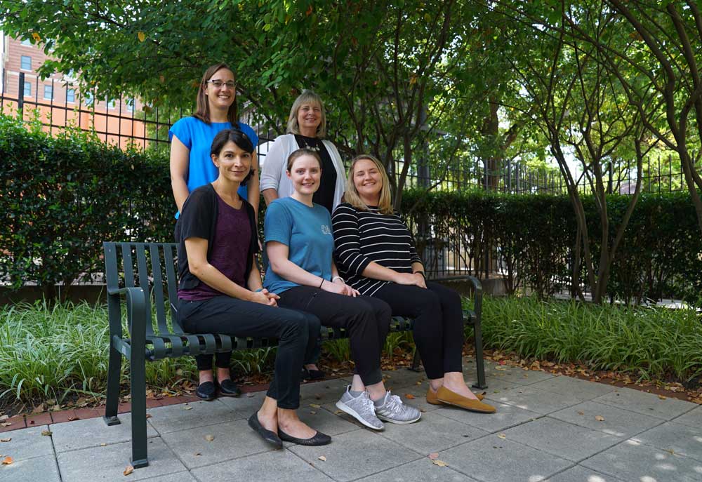 Four women pose outside, three sitting on a bench, and two standing behind the bench. All are smiling.