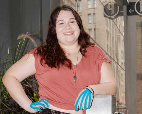 A young woman stands in an urban environment. She is smiling and appears confident. She has blue therapy tape on the back of each hand. The tape strips run from each fingertip to her wrist.