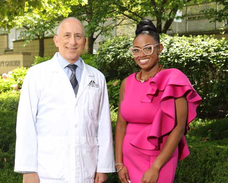 A man in a white doctor’s coat and a young woman stand outside in Kennedy Krieger’s therapy garden. Both are smiling.