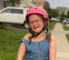 A young girl rides a scooter down a sidewalk. She is wearing a pink helmet. 