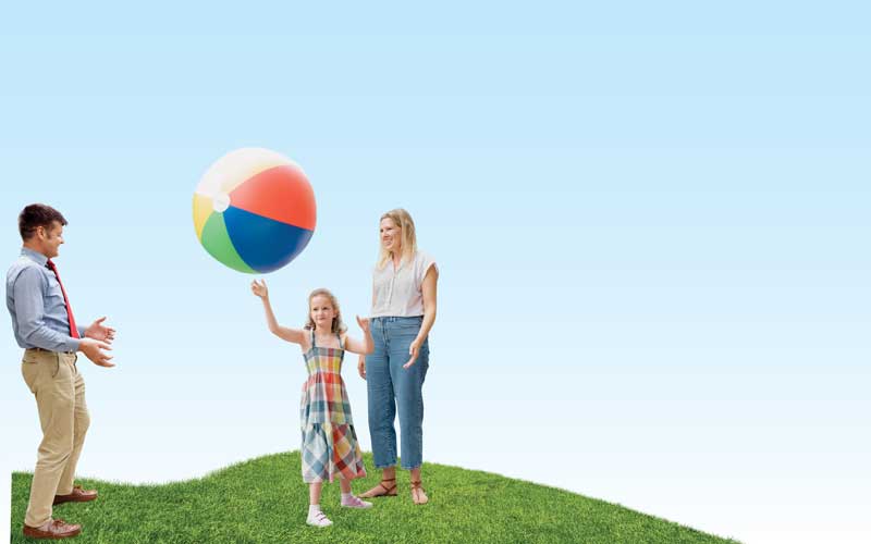 A young girl tosses a large inflatable beach ball outside with a man and a woman. The adults are smiling. The girl concentrates on throwing the ball to the man.