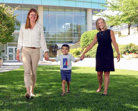 A young boy stands outside between two women, who each hold one of his hands. Everyone is smiling. The boy is wearing glasses and a kippa and has a birthmark on his right ear and on the right side of his neck. There is a Kennedy Krieger building behind them.