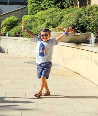 A young boy stands outside with his hands held high, and a big smile on his face. He wears glasses and a kippa, and has a birthmark on his right ear and on the right side of his neck.