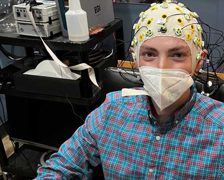 A young man sits in chair in a clinical room. He wears a face mask and a snug-fitting EEG cap fitted with dozens of electrodes.