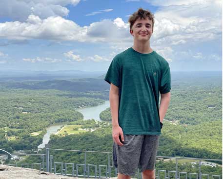 A young man smiles while standing atop a hill or summit. Behind him is a beautiful landscape, with trees, mountains and a river. 