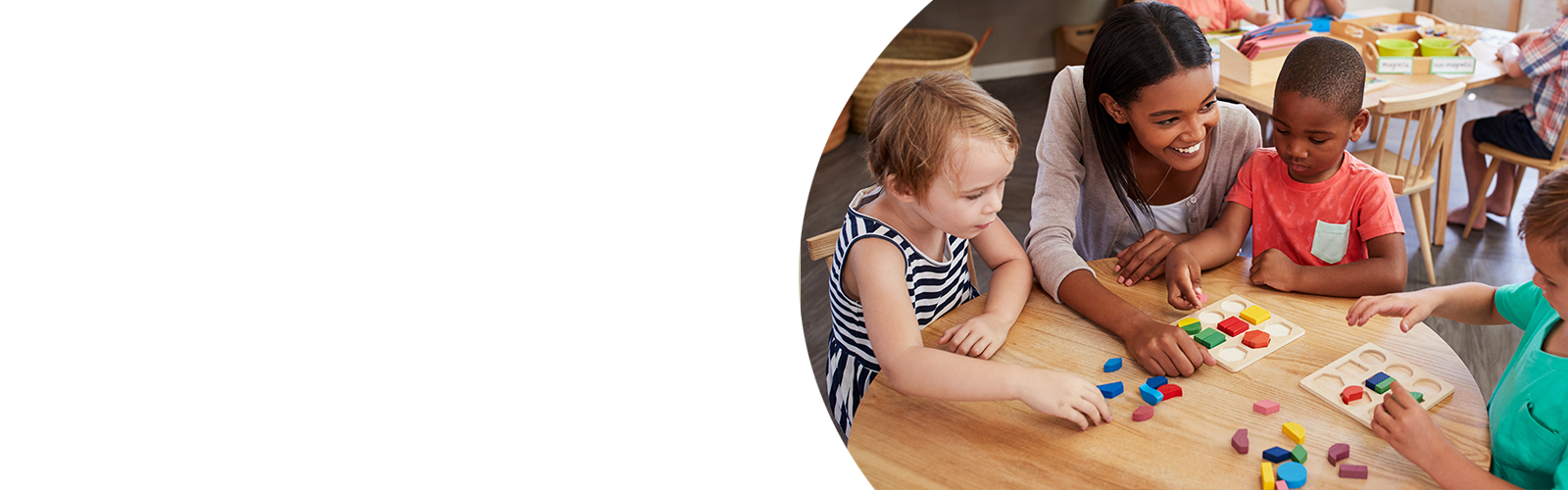 A teacher smiles as she helps three pre-school students with an activity at their desk.