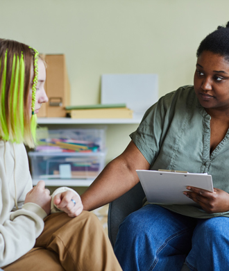 A teenage girl speaks to a social worker, who is sitting to her right. The social worker is listening intently and holding a clipboard.