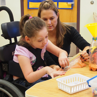A young girl in a wheelchair does a therapeutic activity at a table, while her therapist--an adult woman--sits immediately to her left. A water bottle with a yellow funnel sits on the table.