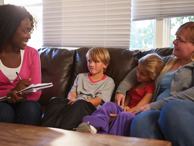 A social worker visits a mother and her two kids at home. All four are sitting on a sofa, with the social worker smiling as she holds a notepad