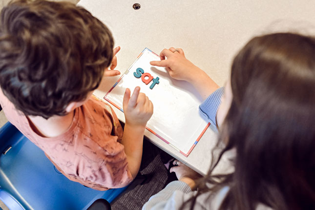 A SLP and patient spell out words on a small tabletop white board with bright letters.