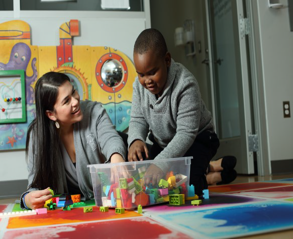 A child plays with blocks while a therapist leans next to them and smiles.