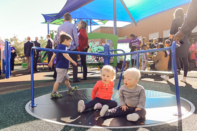 An event at World of Care's playground. In the foreground of the photo are two toddlers sitting on a sit and spin.