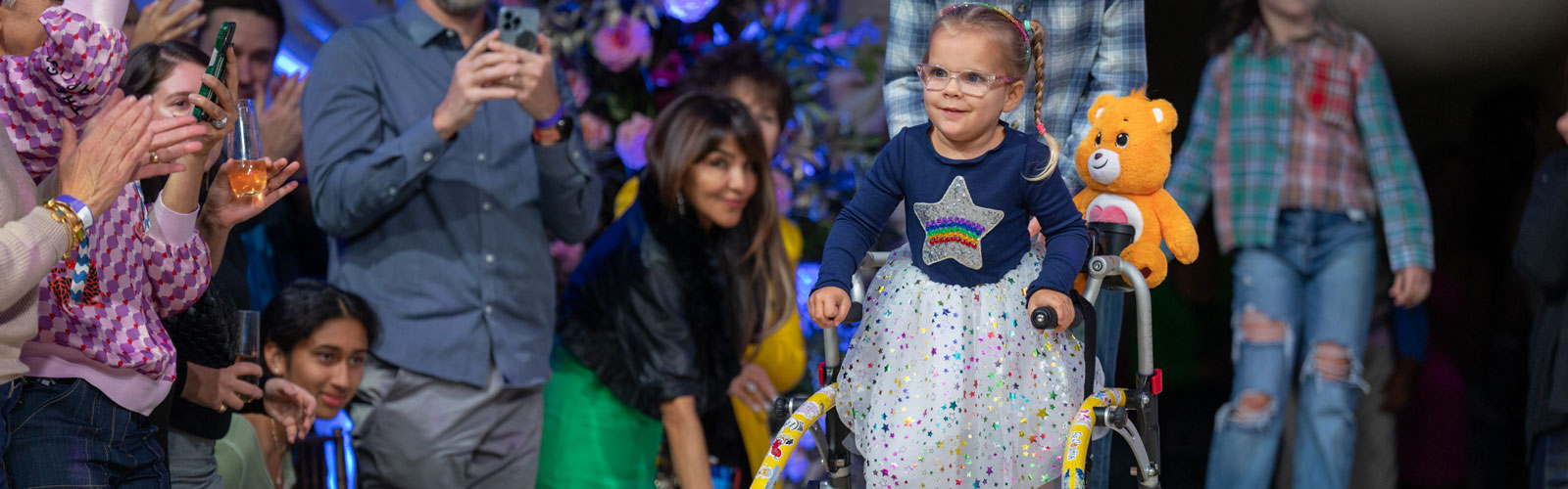 A young girl using a walker walks down a runway while wearing a blue sweater with a  star that has a rainbow in the middle, with a polka dot tutu.