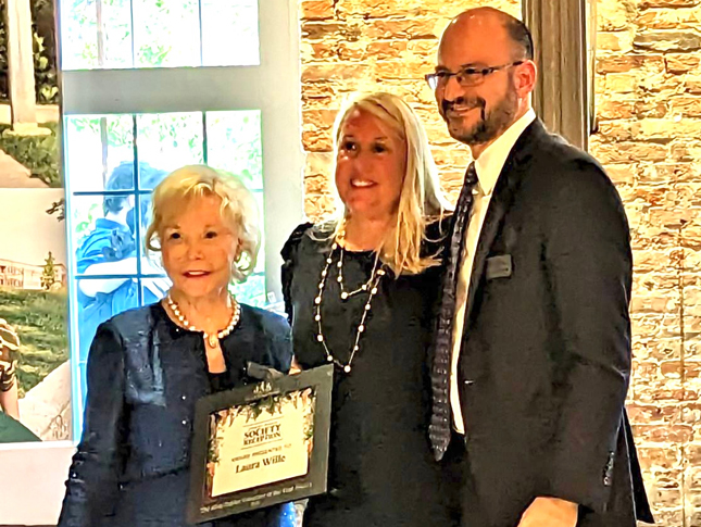 Laura Wille receives the Mimi Baklor Volunteer of the Year Award. She is standing in the center, with a woman wearing a blue blazer and pearls to her left and a man in a suit to the right.