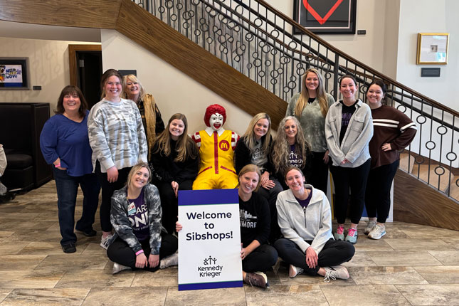 A group of people pose for a photo around a statue of Ronald McDonald sitting on a bench at Ronald McDonald House. A person sitting in front of the statue is holding up a Kennedy Krieger branded sign that says Welcome to Sibshops!