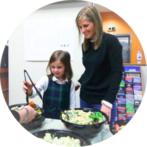 A young girl uses tongs to filler her plate during a WIN inpatient dinner. A woman stands over her shoulder and smiles