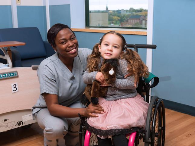 A nurse kneels next to a young girl in a wheelchair for a posed photo. She and the young girl are both smiling.