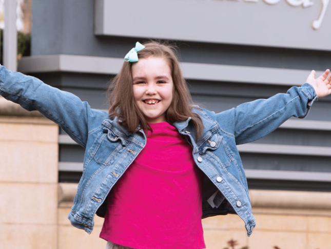 A young girl smiles with her arms outstretched. She has faint port wine-colored birthmarks across her forehead.