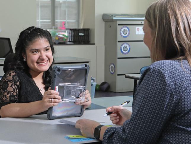 A Kennedy Krieger student smiles while sitting across a desk from her teacher.
