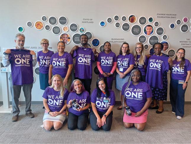 A group of Kennedy Krieger employees smiling as they wear or hold employee giving t-shirts. The purple shirts have We Are One in large white font, with Kennedy Krieger underneath in smaller font.