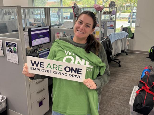 A Kennedy Krieger smiles while standing near her workstation and holding a We Are On Employee Giving Drive sign.