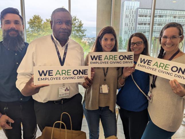 Five Kennedy Krieger staff members smile in a conference room, with three of them holding We Are One Employee Giving Drive signs.
