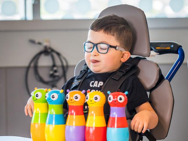 A little boy sitting in a mobility device smiles while playing with toys.