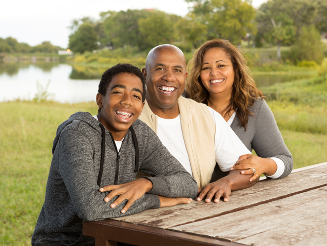 A teenage boy smiles while sitting at a picnic table with his parents.