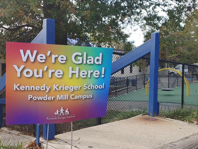 A colorful yard sign sits outside the playground at Power Mill Campus. The sign says We're glad you're here in white letters, with the campus name and Kennedy Krieger logo beneath that message.