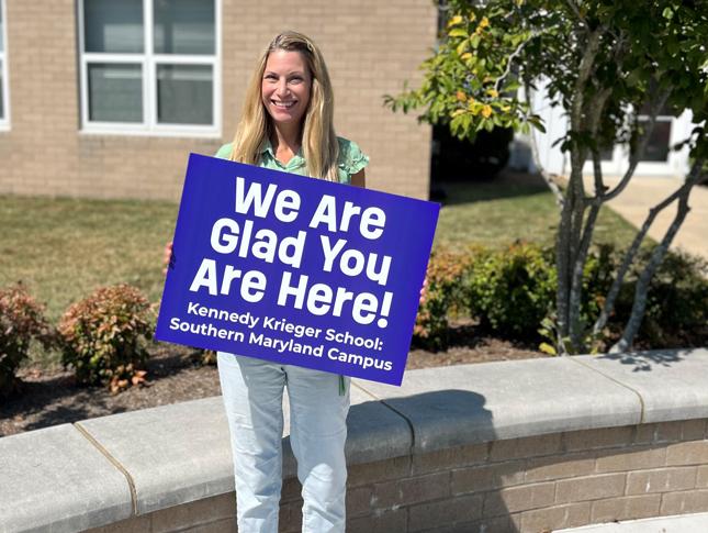 A woman stands outside the Southern Maryland Campus building holding a sign that says We are glad you are here! Kennedy Krieger School: Southern Maryland Campus.