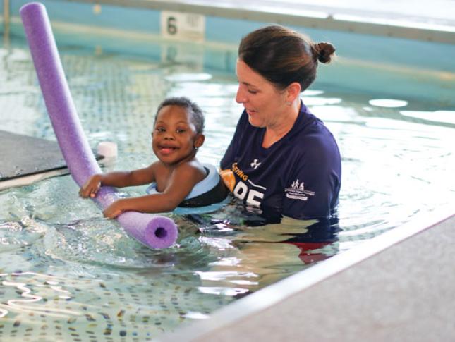 A young boy wearing a floatation device in a pool is held by a female therapist during an aquatic therapy session.