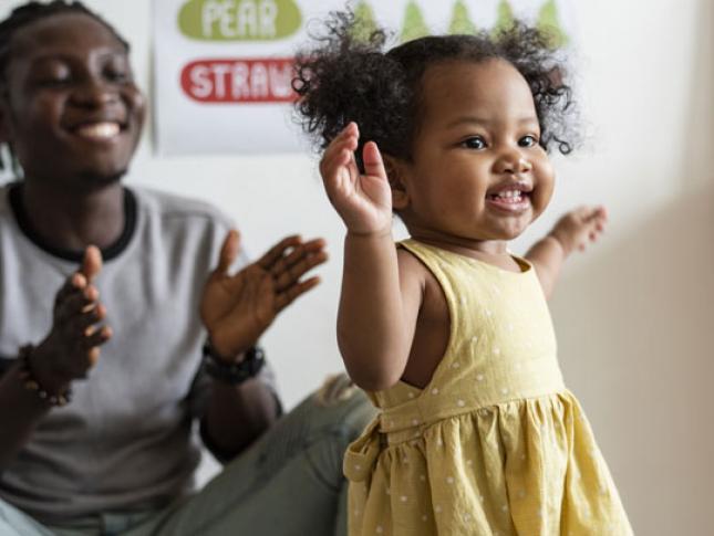 Man in background holds out hands as small girl walks toward camera