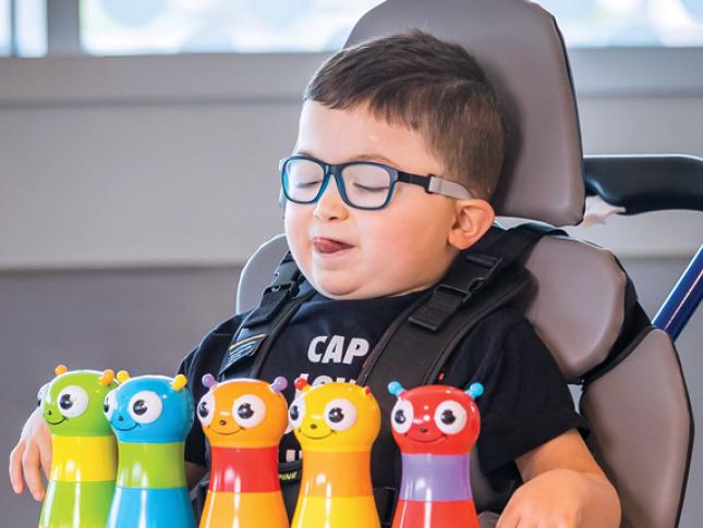 A little boy sitting in a mobility device smiles while playing with toys.
