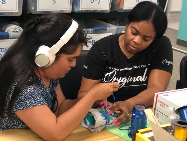 Two young women sit at table and wrap pieces of paper in a blue string. The woman sitting to the left is earing white headphones.