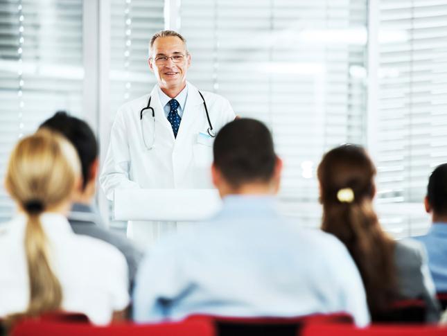 A male doctor wearing a white lab coat gives a seminar inside of a classroom. The backs of give trainee's heads can be seen in the foreground.