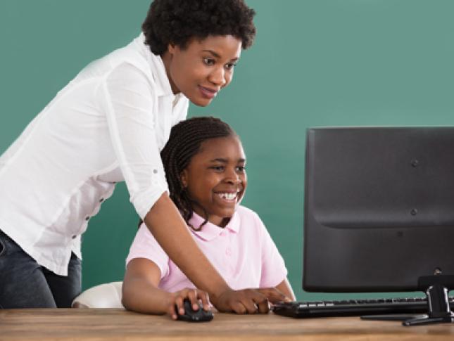 A woman stands and assists a little girl as the girl sits at a desk and uses a computer.