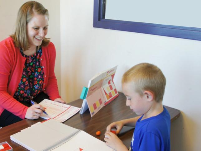 A neuropsychologist smiles during an appointment with her patient, a young boy that is sitting on the other side of a desk.