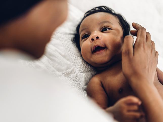 A newborn lays on a bed and looks at its mother. The mother's back is to the camera, with her right hand gently touching the side of the baby's head.