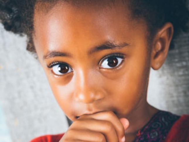 A wide-eyed little girl looks to the camera while resting her fingers against her mouth.