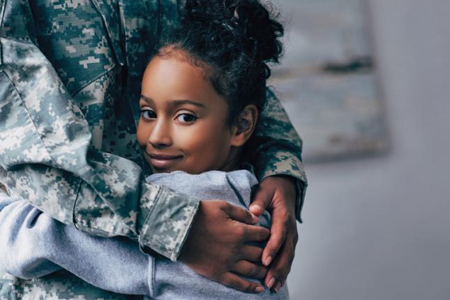 A young girl looks at the camera as she hugs an adult wearing a military uniform