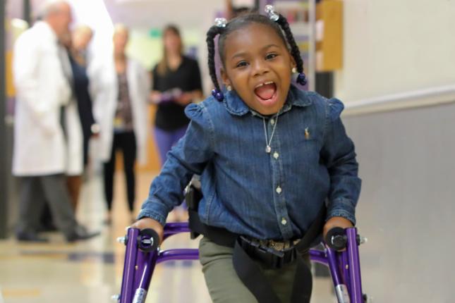 A photo of Journee in the hallway at Kennedy Krieger, smiling at the camera as she walks with the assistance of a walker