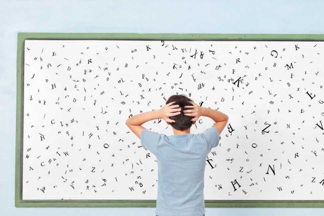 A child stands in front of a board with letters, holding his arms on his head in confusion