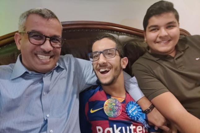 Ali, a father, sits on the sofa with his two adult sons.