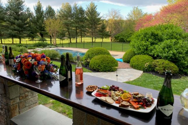 Food and wine sit laid out on a table outside, with rows of colorful trees and nature in the background.