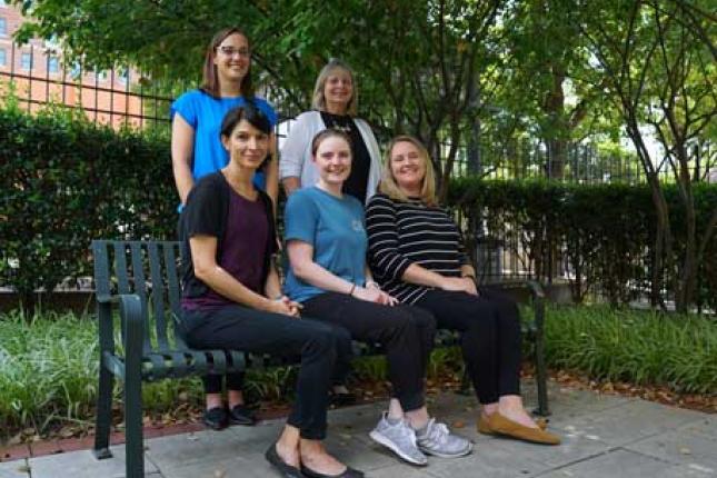 Four women pose outside, three sitting on a bench, and two standing behind the bench. All are smiling.