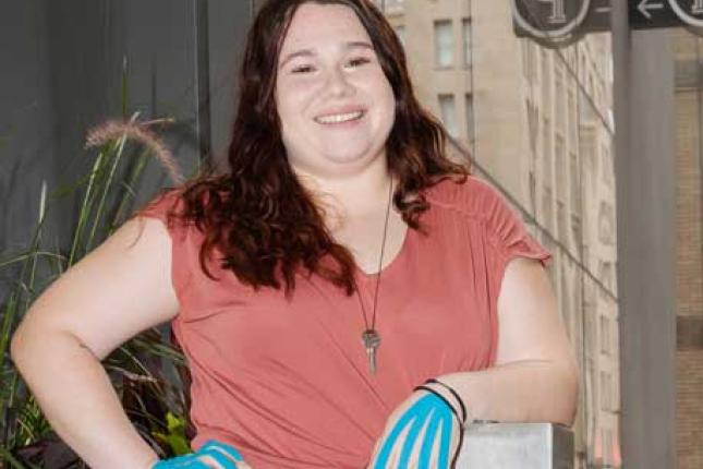 A young woman stands in an urban environment. She is smiling and appears confident. She has blue therapy tape on the back of each hand. The tape strips run from each fingertip to her wrist.